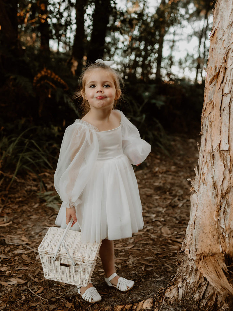 A flower girl stands holding a basket, wearing our Eva tulle sleeve dress in light ivory.