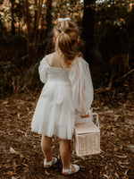 The back of our Eva flower girl dress, worn by a young flower girl. She holds a basket. Showing the back zip closure and tulle bow at the waist.