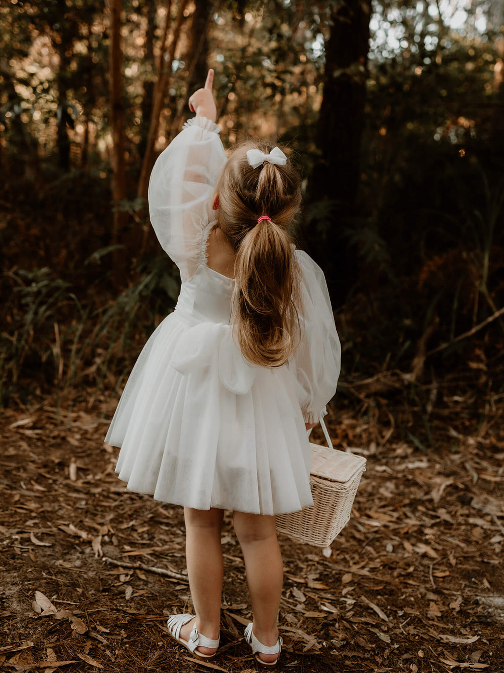 Eva tulle sleeve flower girl dress in ivory is worn by a young girl standing on a bridge. She wears a small ivory tulle bow in her hair, and stands with a flower girl basket at her feet.