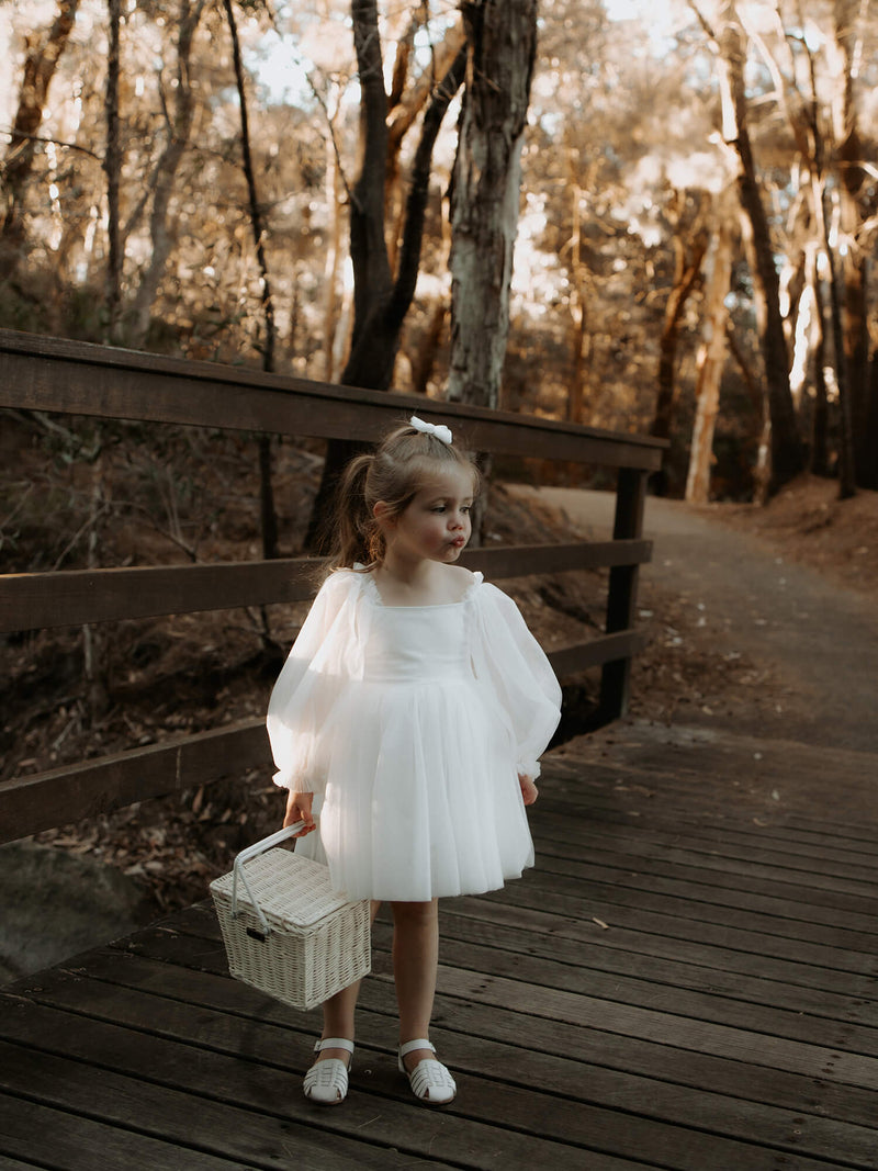 Flower girl stands on a bridge in the forest wearing our Eva tulle sleeve flower girl dress in ivory. She holds a basket and wears a small tulle bow in her hair.