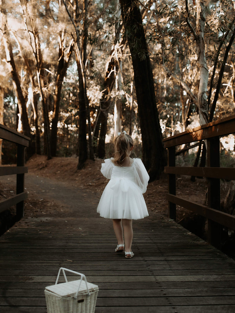 Eva tulle sleeve flower girl dress in ivory is worn by a young girl, she is walking over a bridge in the forest, showing the back of the dress, with a zip closure and tulle bow tied at the waist.