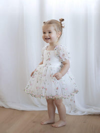 Child wearing our Fleur floral flower girl dress standing against a white curtain.