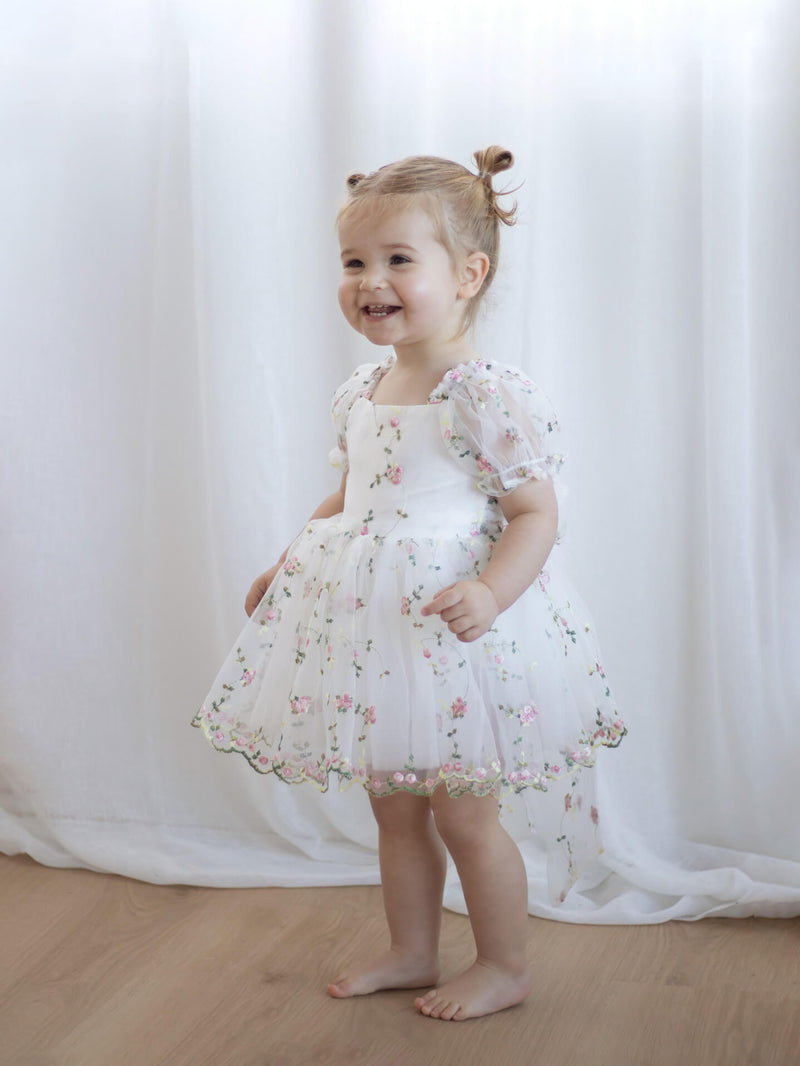 Child wearing our Fleur floral flower girl dress standing against a white curtain.