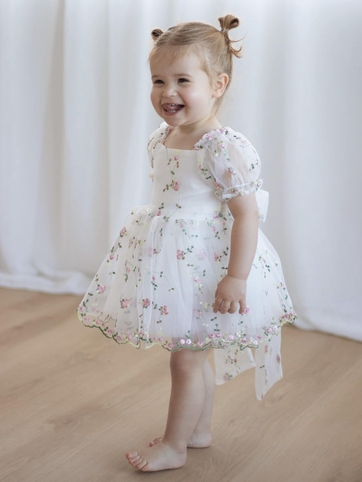 A toddler smiles wearing our Fleur pink floral organza flower girl romper dress, with puff sleeves.