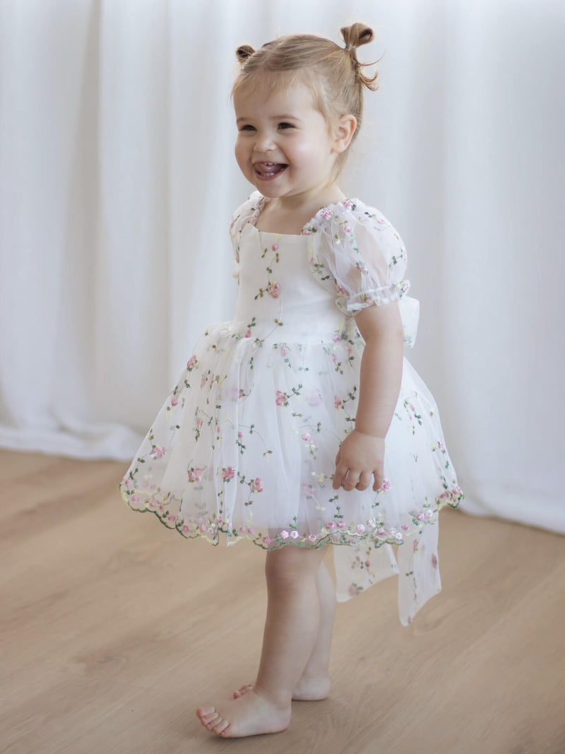 A toddler smiles wearing our Fleur pink floral organza flower girl romper dress, with puff sleeves.