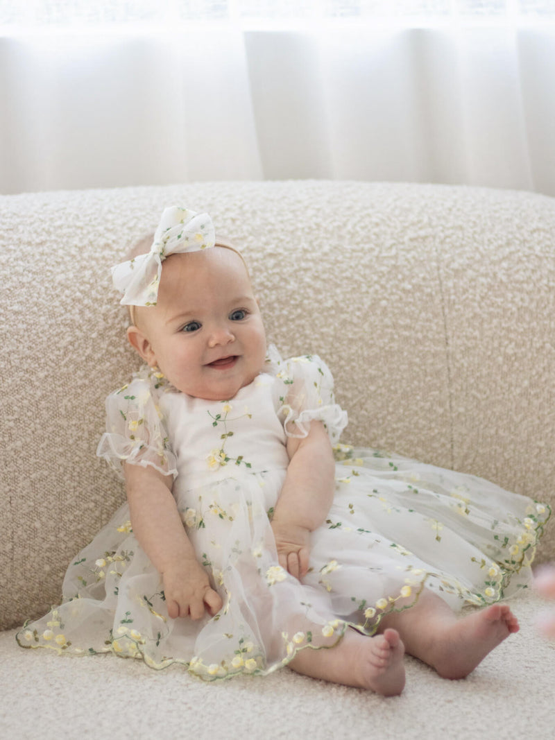 A baby smiles wearing our Fleur lemon blossom baby flower girl romper and matching bow headband.