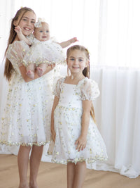 Sisters stand wearing matching Fleur lemon blossom dresses. The oldest sister holds her baby sister, who wears the Fleur lemon blossom baby flower girl romper.