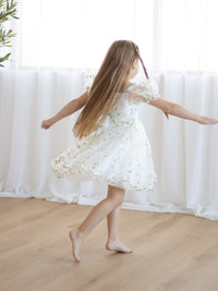 Young girl in a white and lemon yellow floral dress dancing in front of white curtains.