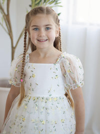 A young girl smiles wearing our Fleur lemon floral embroidered flower girl dress, with puff sleeves.