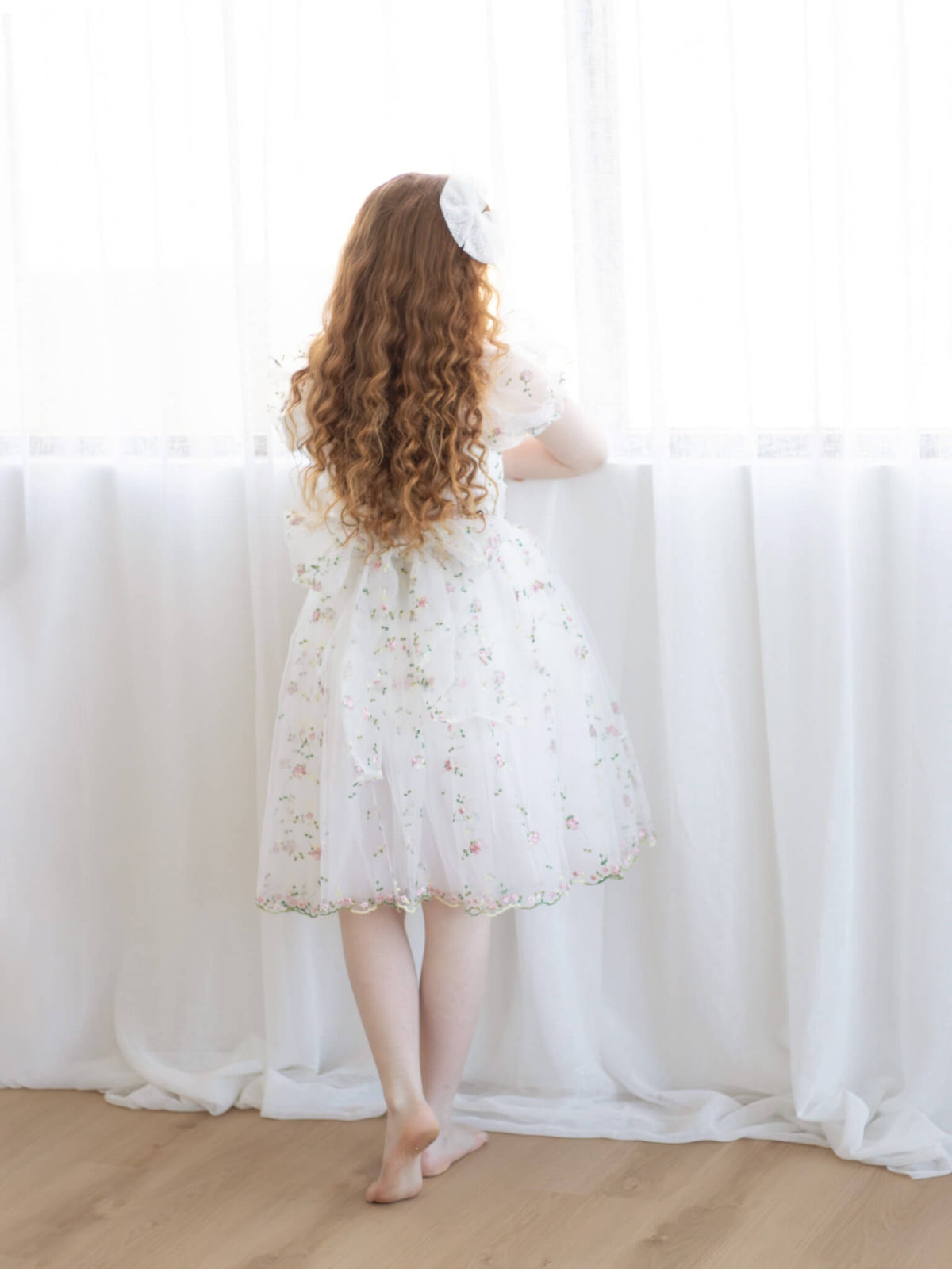A girl stands looking out a window, wearing a pink floral embroidered flower girl dress.