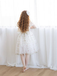 A girl stands looking out a window, wearing a pink floral embroidered flower girl dress.