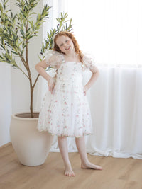 A young girl stands smiling, wearing our Fleur floral organza flower girl dress.