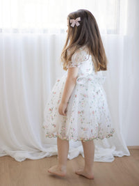 Young girl in a white floral flower girl dress standing in front of sheer curtains.