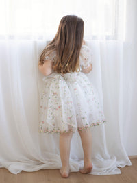 A little girl looks out  a window wearing our Fleur flower girl dress, showing the organza bow at the back.
