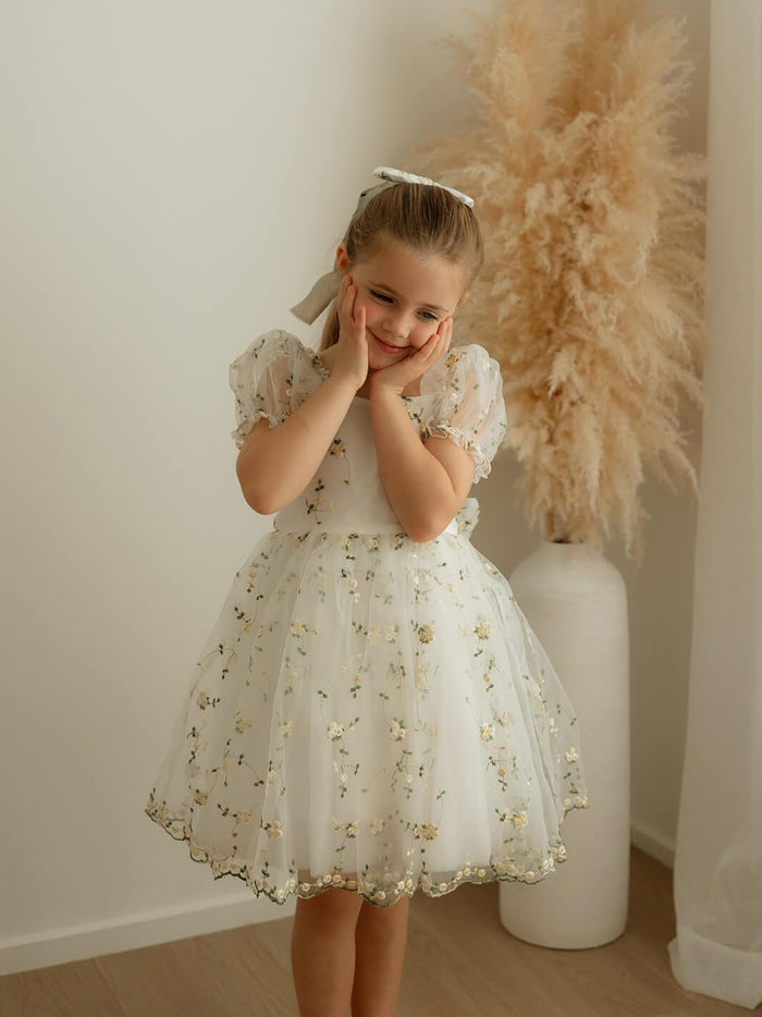 A little girl stands smiling, wearing our Fleur lemon floral flower girl dress.