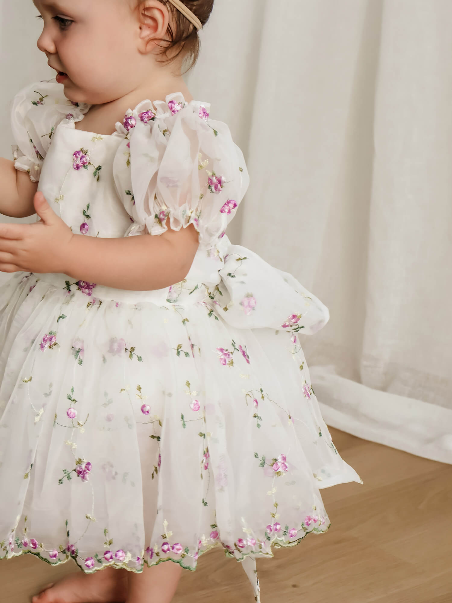 A toddler wearing a white organza puff sleeve flower girl dress with lilac floral embroidery, standing indoors against a soft cream curtain background.