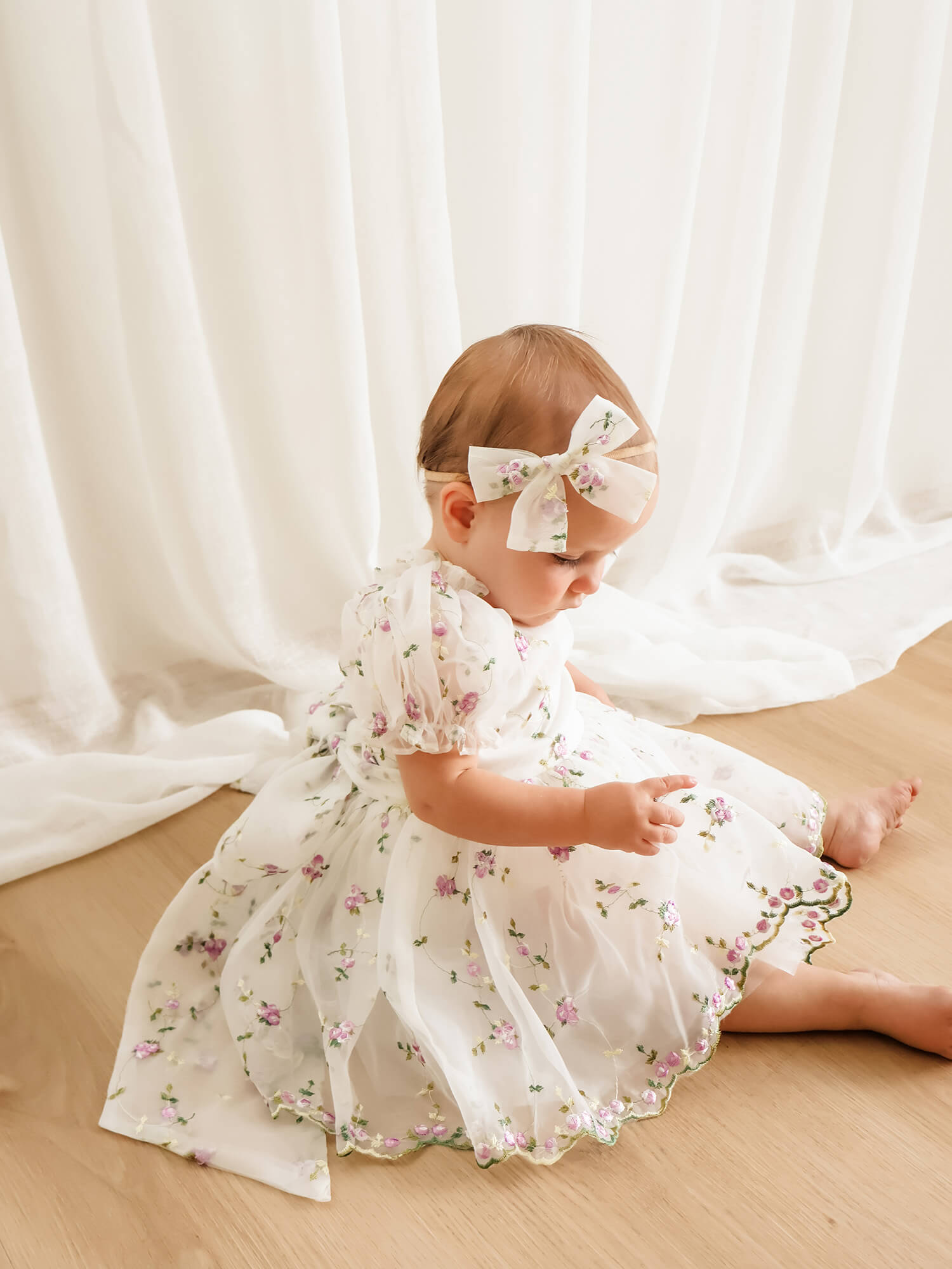 Baby girl sits wearing our Fleur baby flower girl romper, made with ivory and lilac purple floral organza. She also wears a matching organza bow headband.