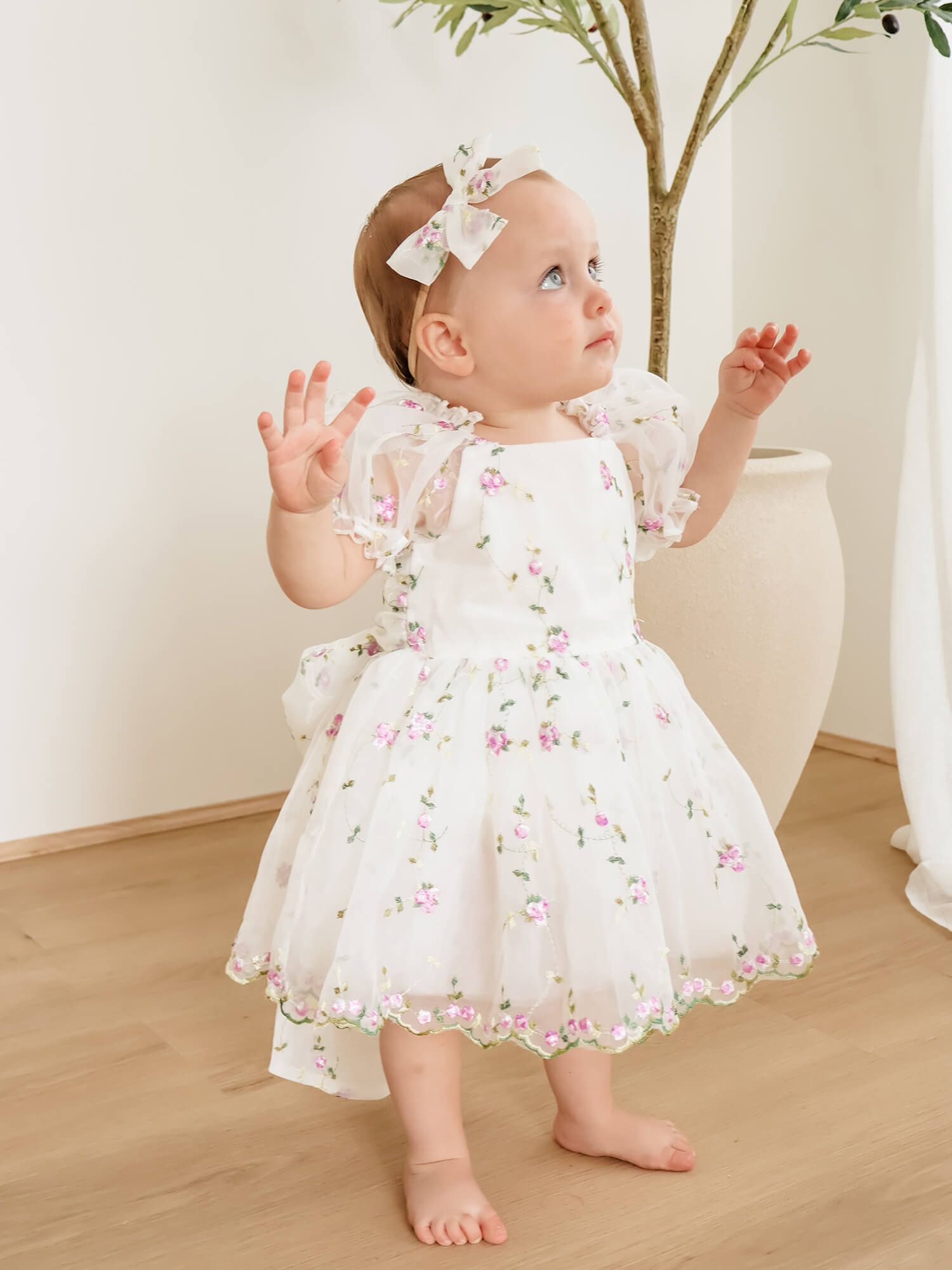 A baby girl wears our Fleur lilac flower girl dress and a matching bow headband, standing in a bright indoor setting.