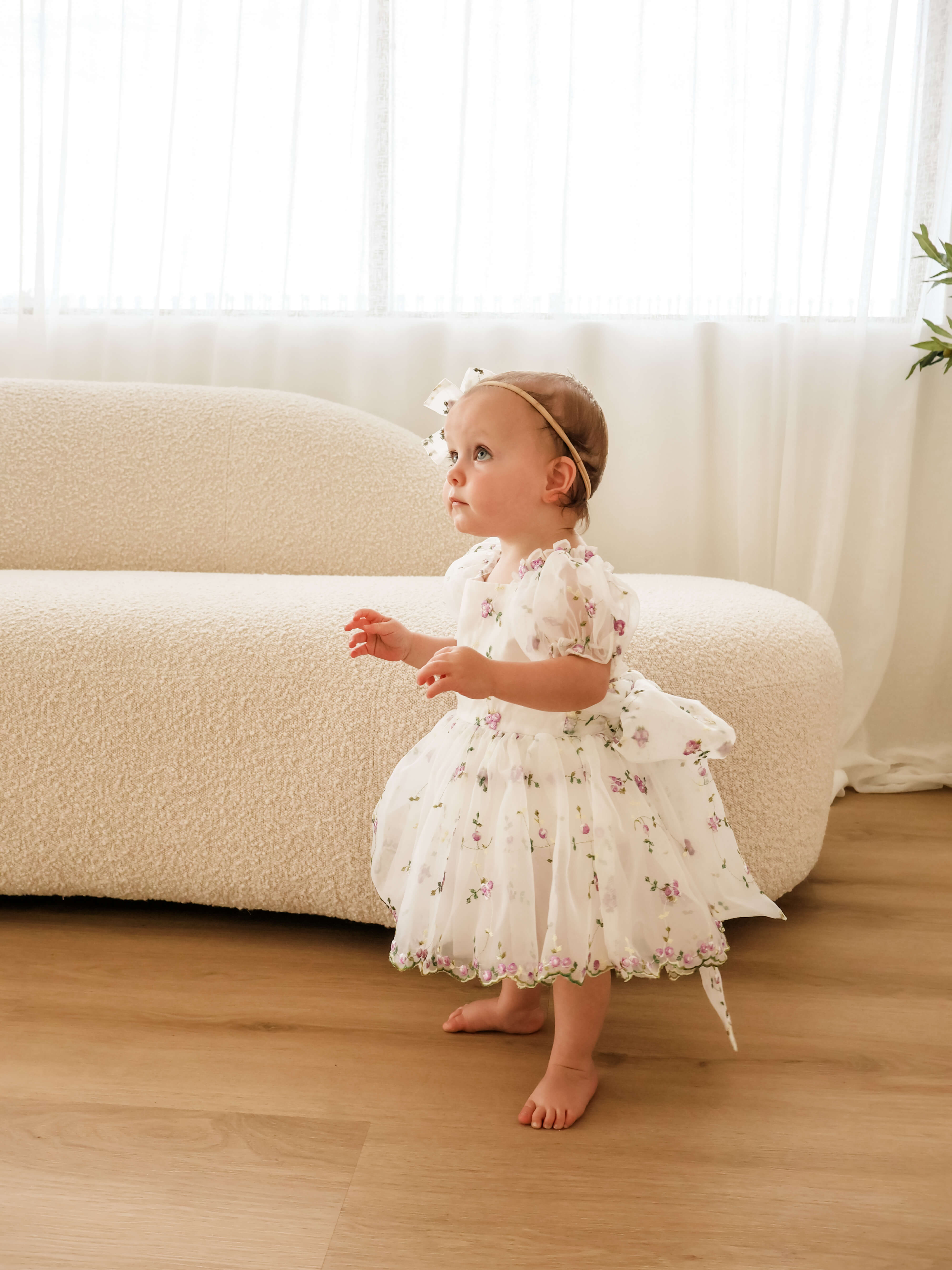 A toddler girl wearing a white and lilac floral baby flower girl dress and matching headband stands barefoot on a light wood floor in front of a cream boucle sofa.