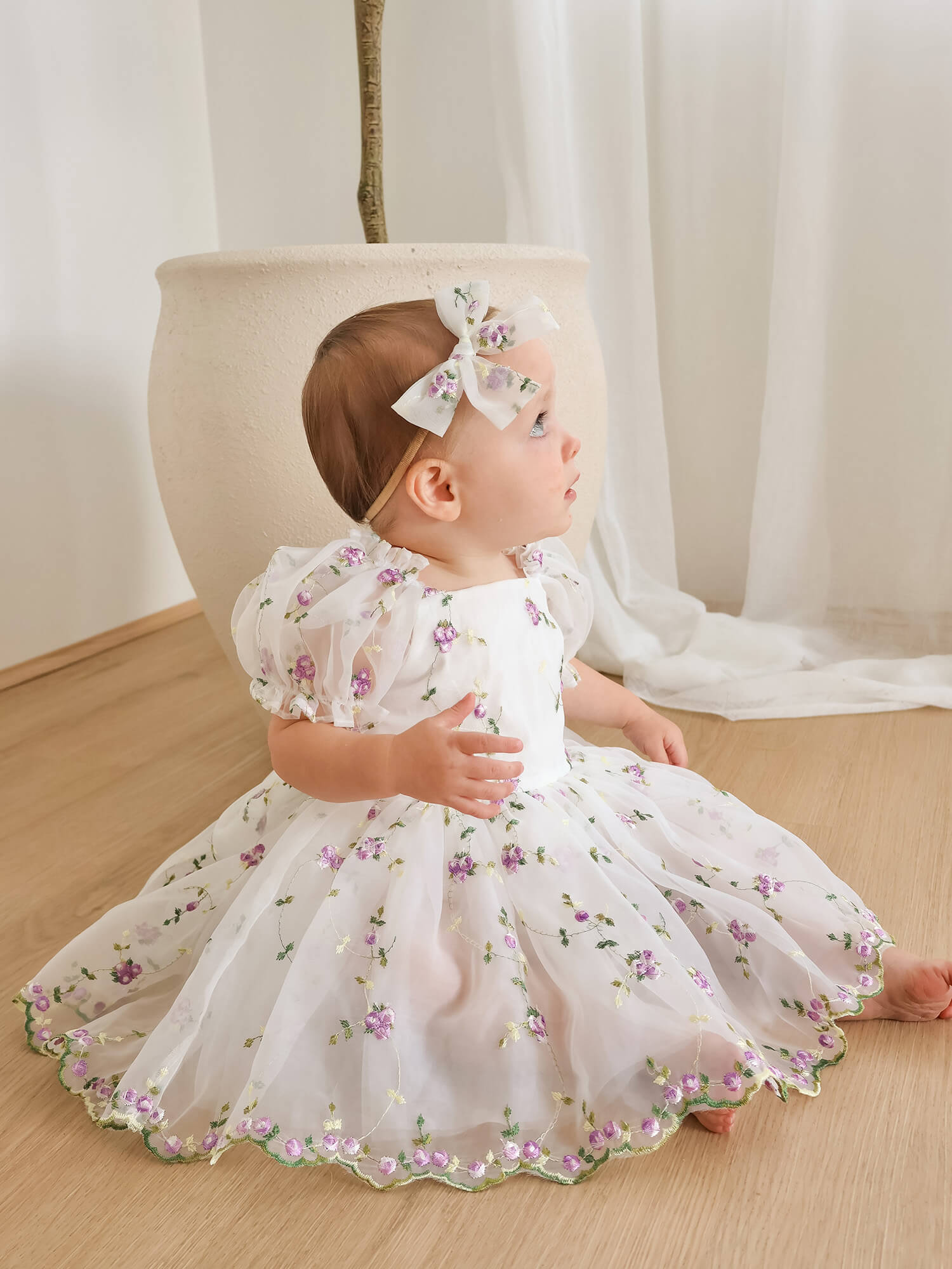 A baby girl wearing a white organza floral first birthday dress with puff sleeves and a matching bow headband, sitting on a light wood floor.