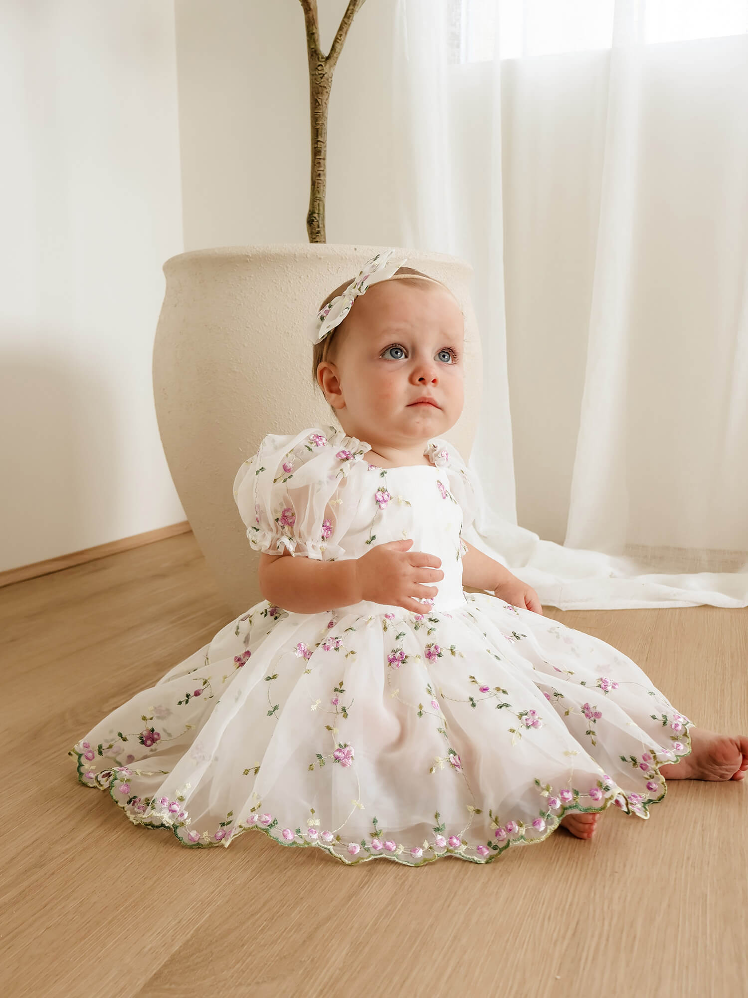 Fleur baby dress in ivory and lilac floral embroidered organza is worn by a baby, she also wears a matching baby bow headband.