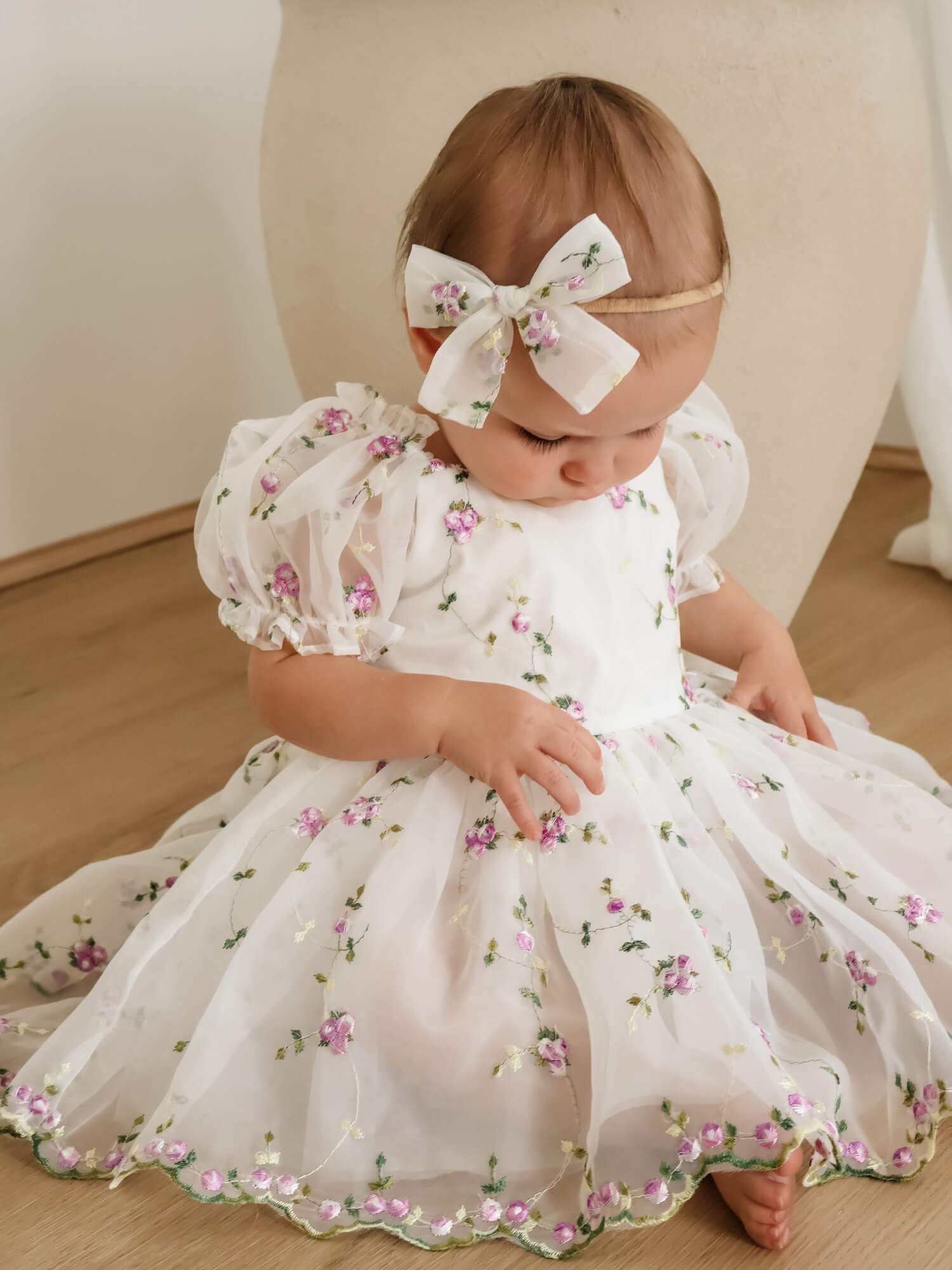 A baby girl wearing a white organza puff sleeve dress with purple floral embroidery and a matching white bow baby headband, sitting on a wooden floor.