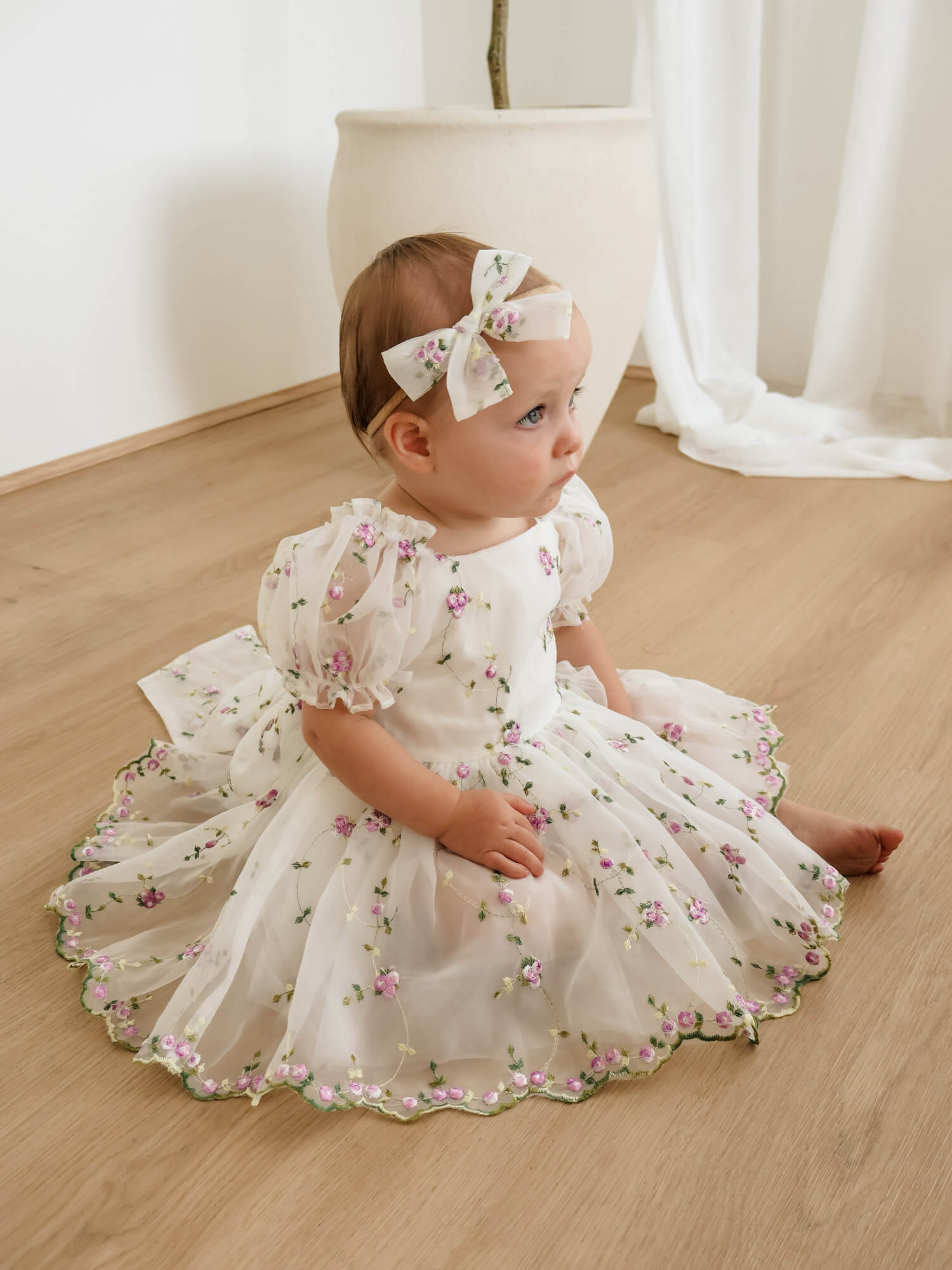 Fleur baby floral flower girl dress is worn by a baby girl, who also wears a matching bow headband.