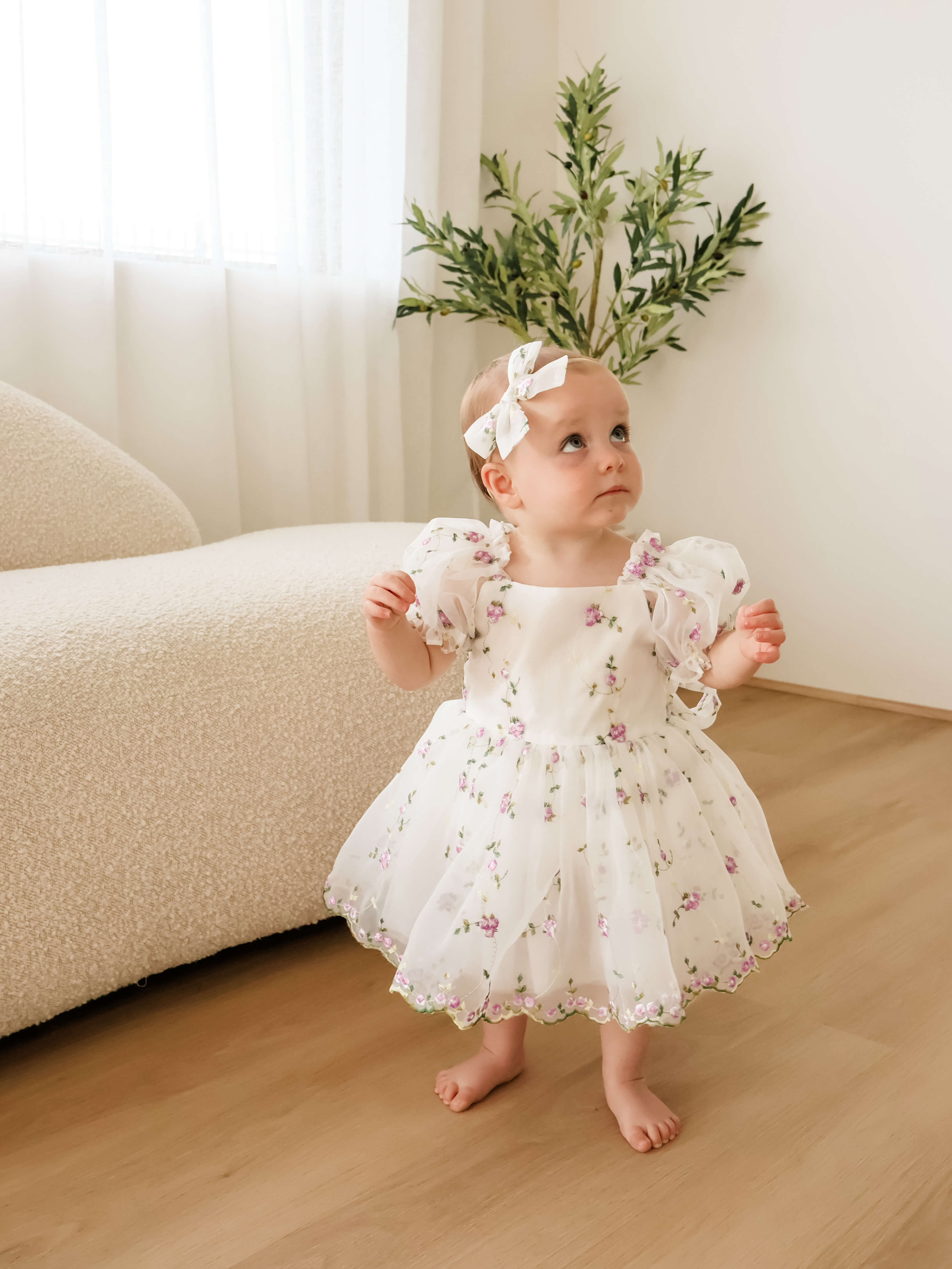 A baby girl wearing a white organza baby flower girl dress with a purple floral print and a matching white bow headband, standing on a wooden floor in a bright room.