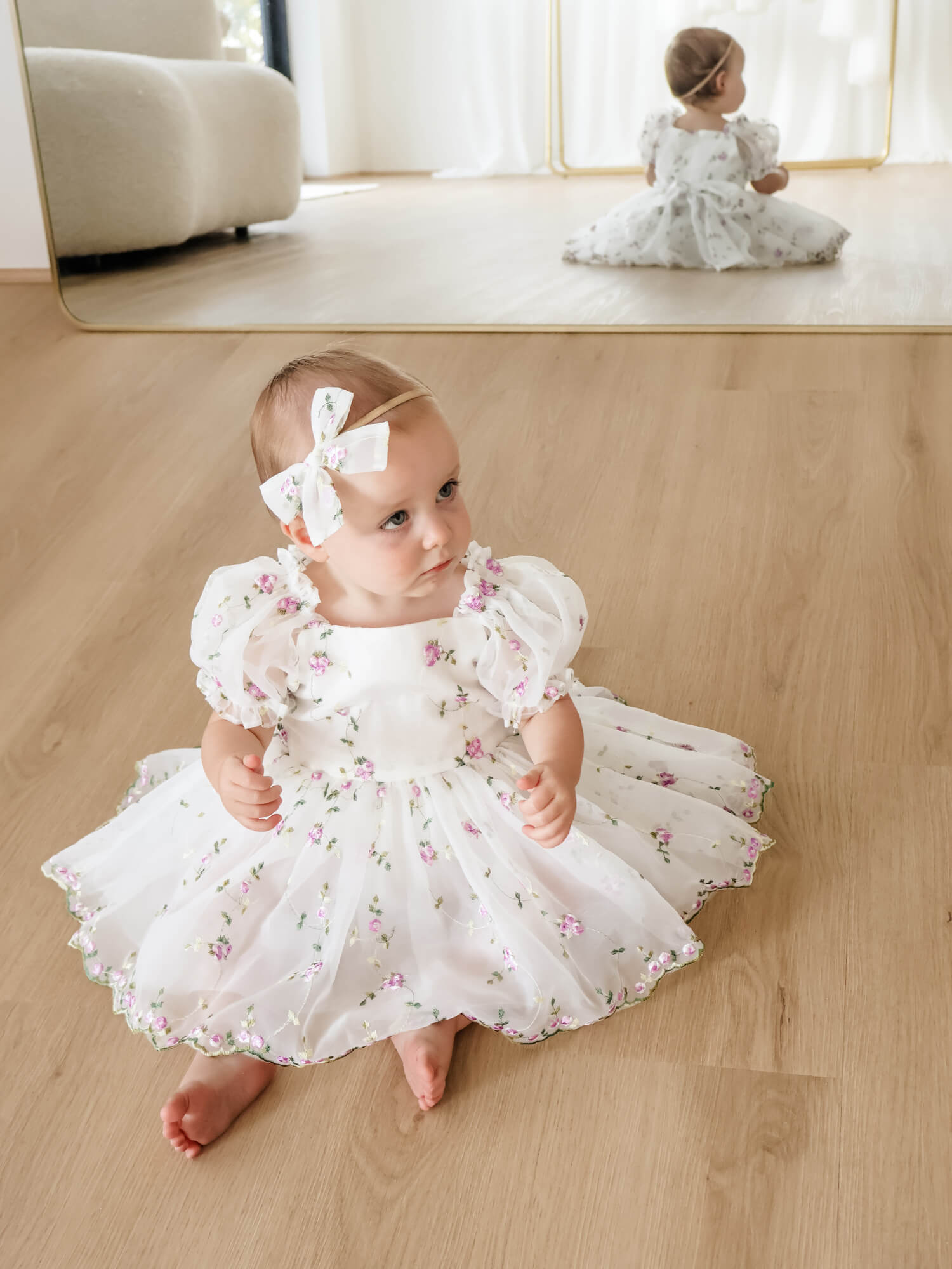 A baby girl sitting on a light wood floor wearing a white puff-sleeve dress with a purple floral print and a matching white bow headband. A large mirror in the background shows her reflection.