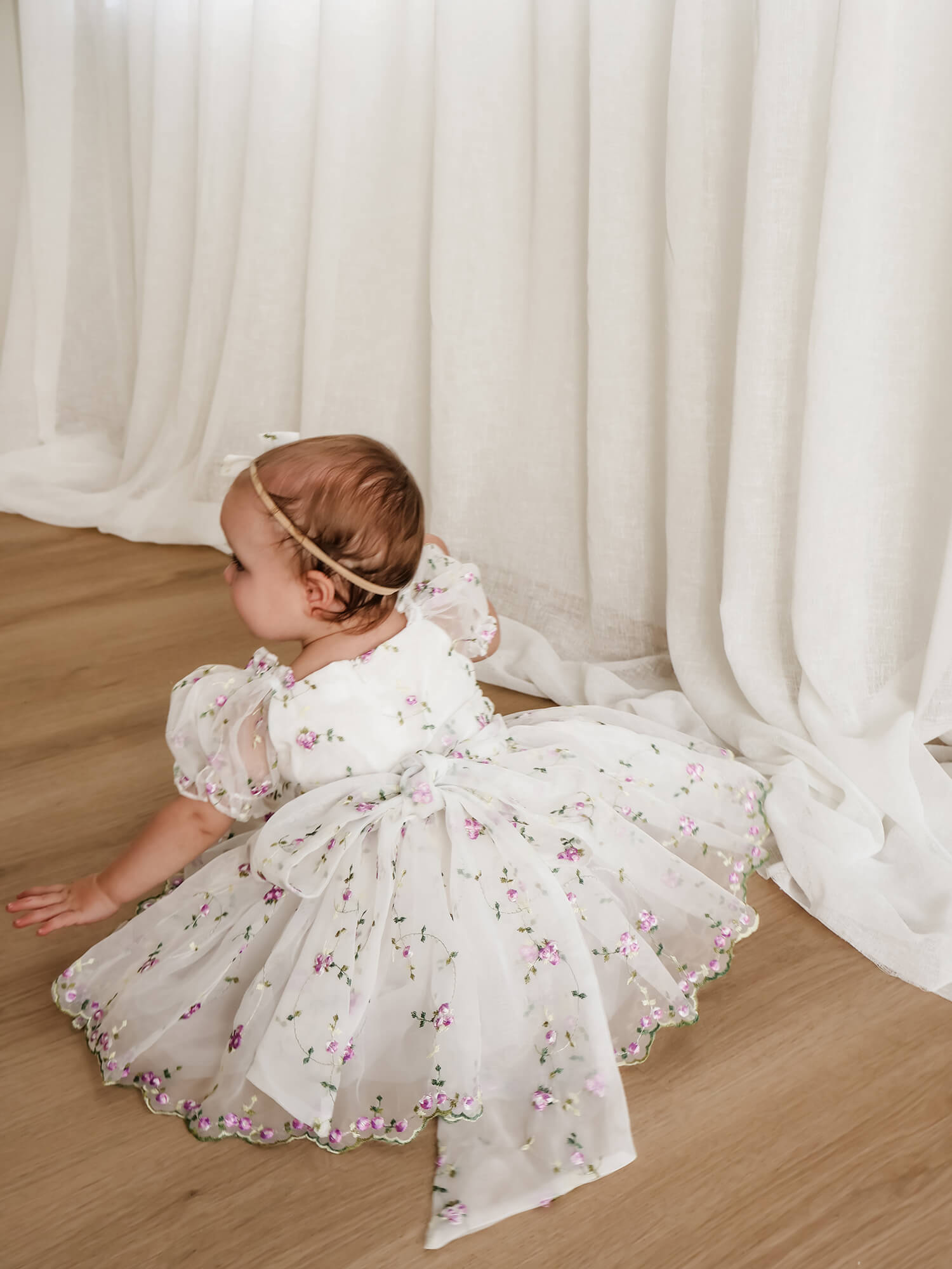 A baby girl wearing a white flower girl dress with lilac purple floral embroidery and a matching headband, sitting on a light wood floor in front of white curtains.
