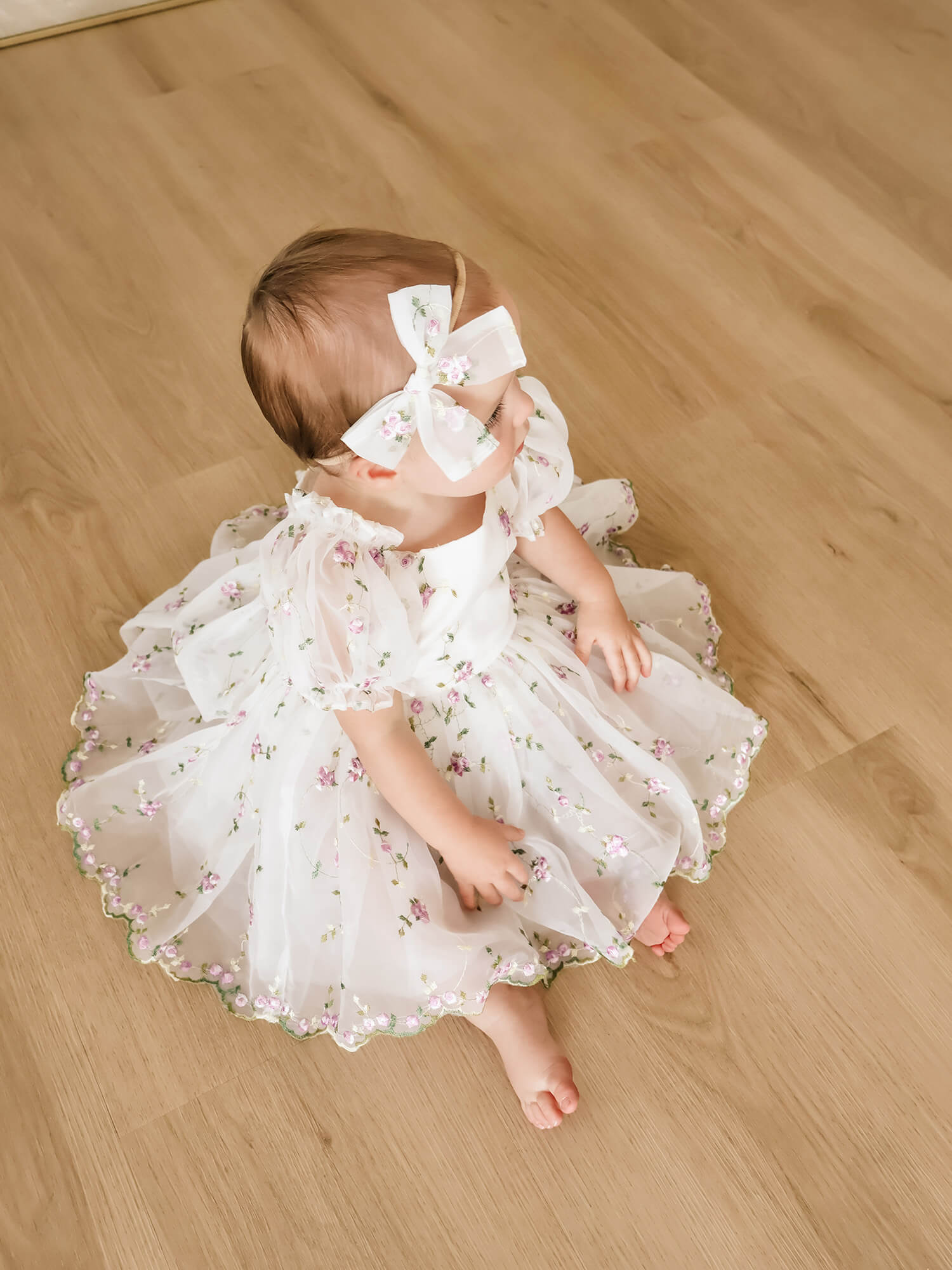 A baby girl sitting on a light wood floor wearing our Fleur lilac baby flower girl dress with purple floral embroidery and a matching white bow headband.
