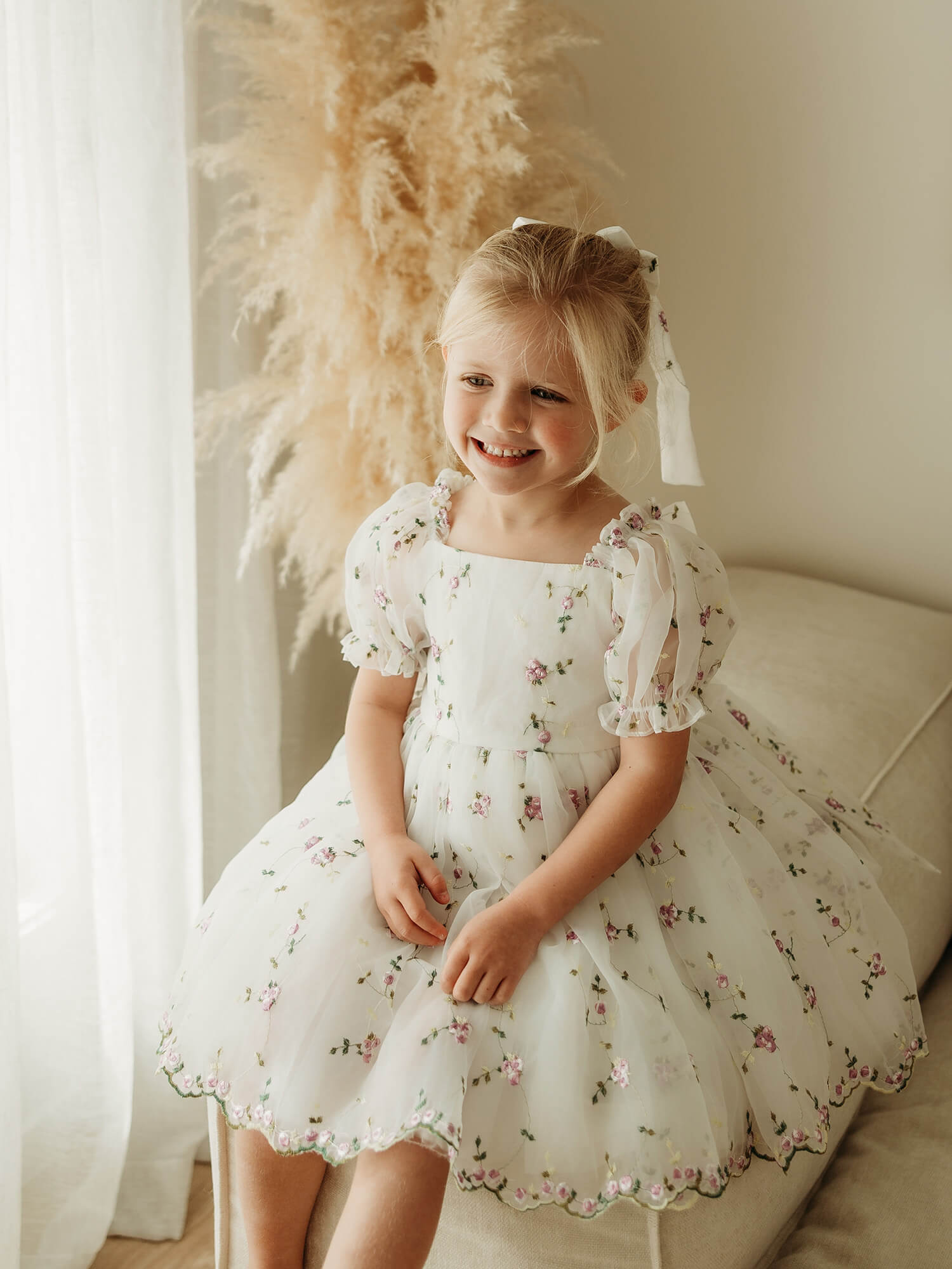 A young girl with blonde hair in a lilac floral bow sits on a beige sofa wearing a white organza flower girl dress with a lilac purple floral print.
