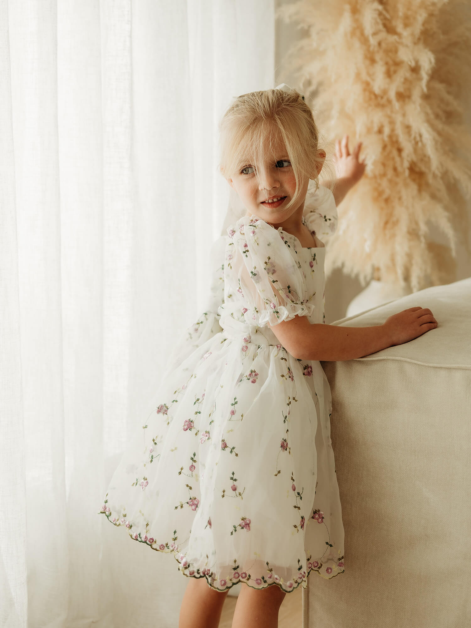 A young blonde girl wearing a white organza flower girl dress with a delicate purple floral print, standing next to a beige sofa in a brightly lit room.