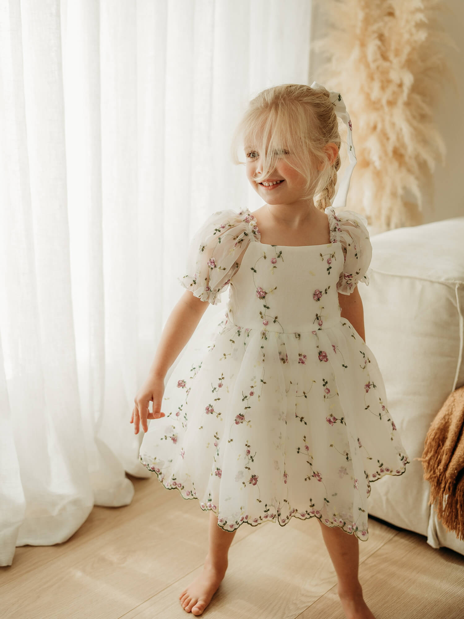 A young girl with blonde hair in a ponytail stands barefoot in a bright room, wearing a white puff-sleeve flower girl dress with a purple floral pattern.