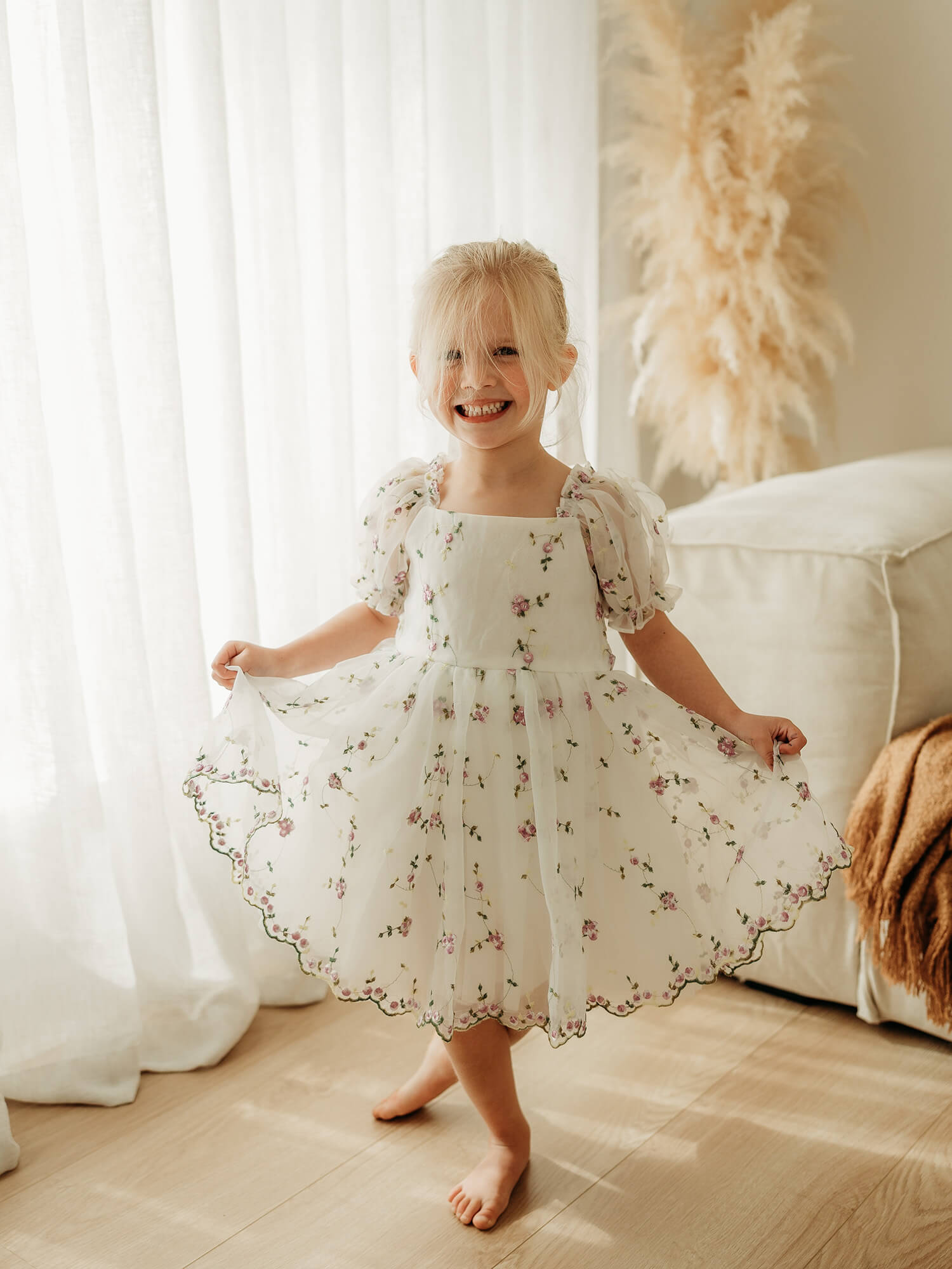 A young blonde girl smiling and twirling in a white organza dress with a delicate purple floral print, standing barefoot on a light wood floor in a bright room.