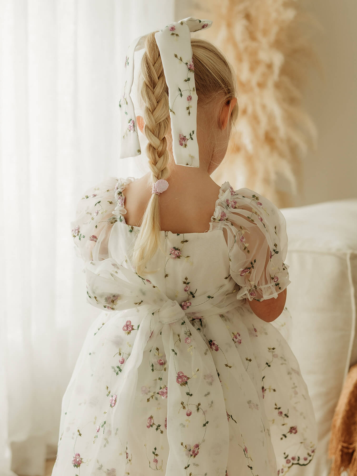 Fleur lilac purple flower girl dress shown from behind on a young girl, showing the puff sleeves, a square neckline, and a large bow at the back, paired with a matching hair bow. 