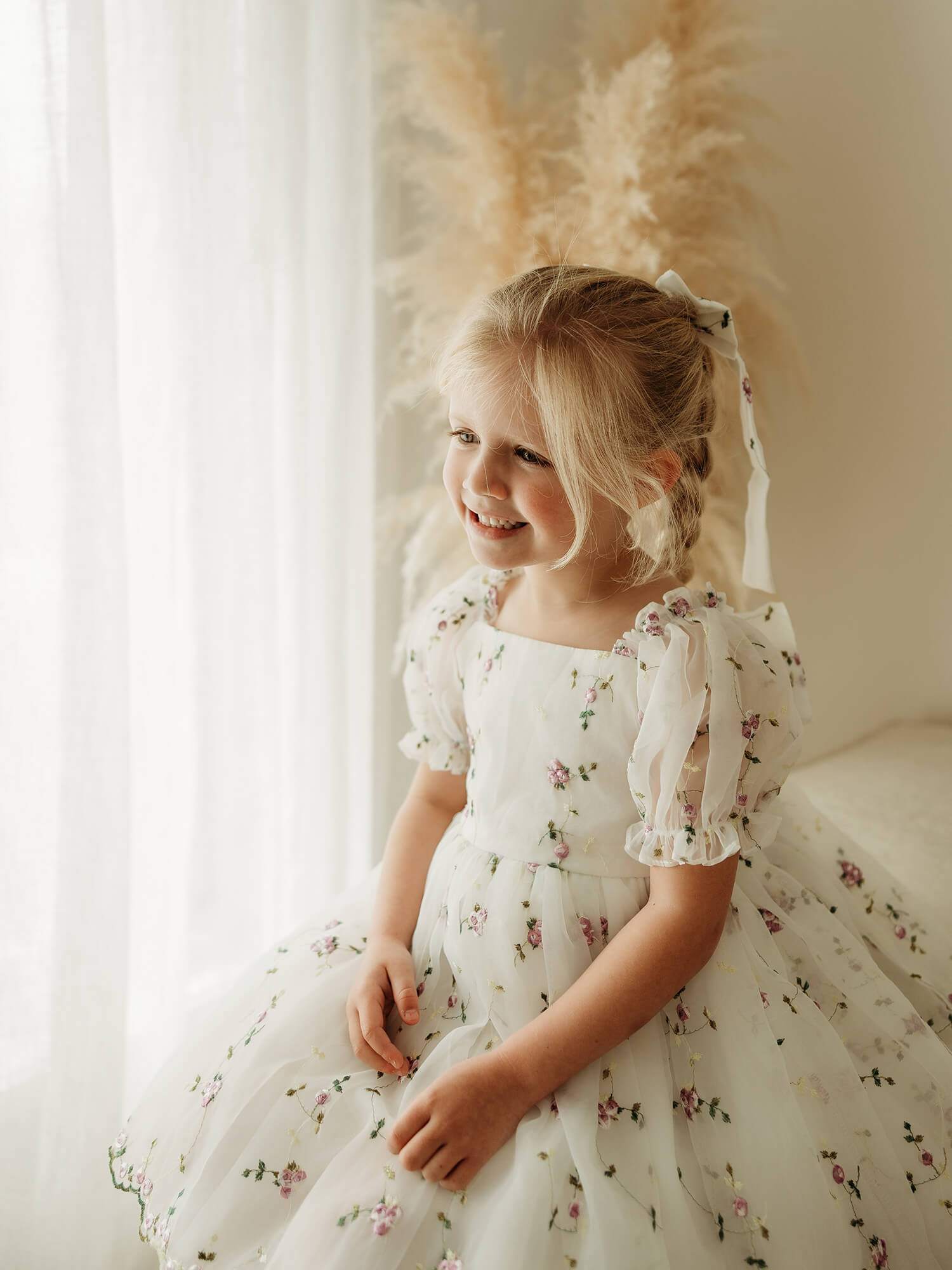 A young blonde girl wearing a white organza flower girl dress with lilac floral embroidery, sitting in a brightly lit room with sheer curtains and pampas grass in the background.