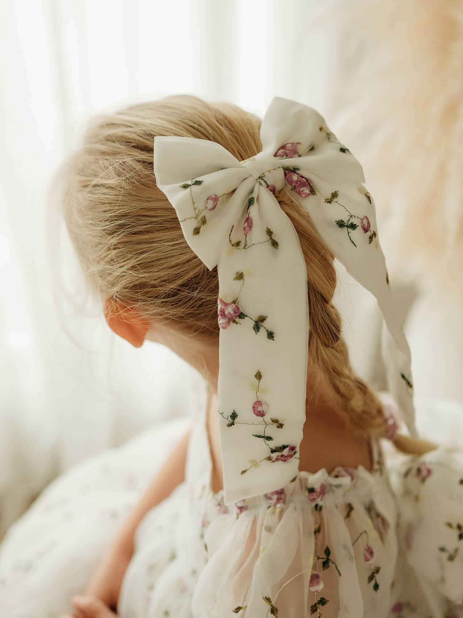 A close-up back view of a young girl wearing a large fabric hair bow with lilac floral embroidery, paired with a matching lilac floral flower girl dress.