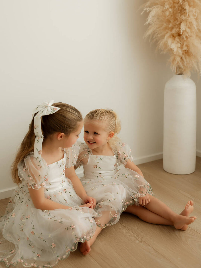 Two young girls sit together wearing matching white floral flower girl dresses.