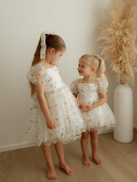 Sisters look at each other wearing matching embroidered organza flower girl outfits.
