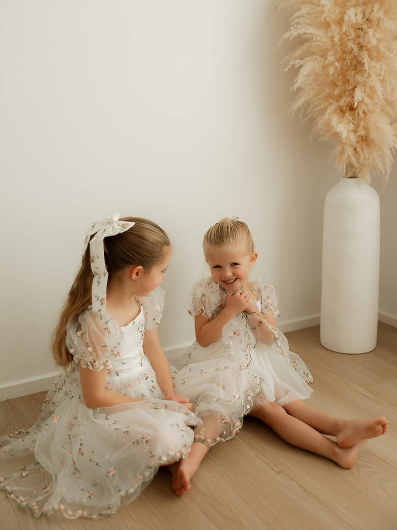 Sisters sit wearing matching Fleur meadow flower girl dress and Fleur meadow flower girl romper.