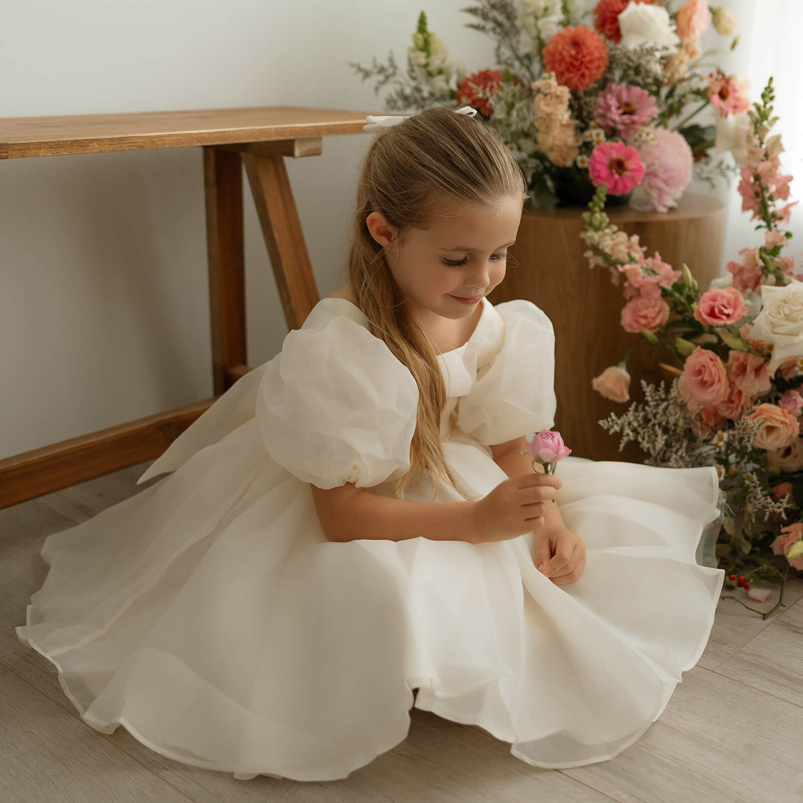 Cream puff sleeve flower girl dress is worn by a young girl who sits amongst flowers.