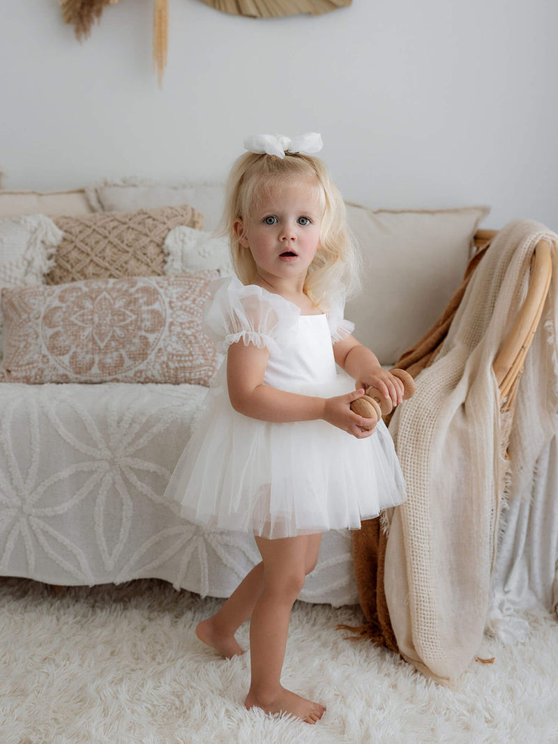 Little girl wearing our cream Gabrielle baby flower girl romper with puff sleeves, holding a wooden toy.