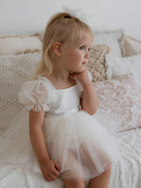 Little girl sitting and looking away, wearing our Gabrielle cream baby flower girl romper dress with puff tulle sleeves.