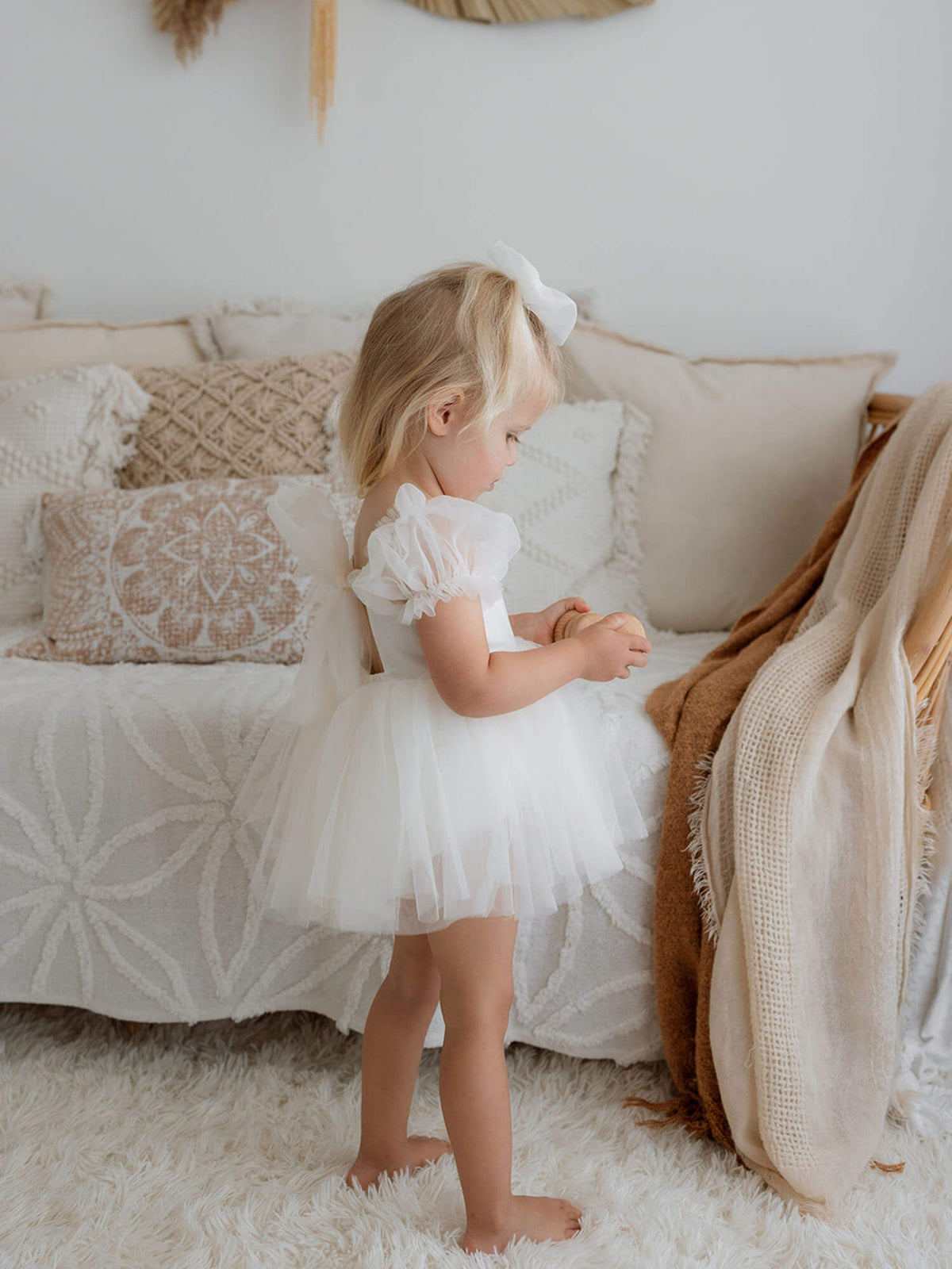 A little girl wearing our cream Gabrielle baby flower girl outfit with a tulle bow back, holding a wooden toy, with a matching tulle bow in her hair.