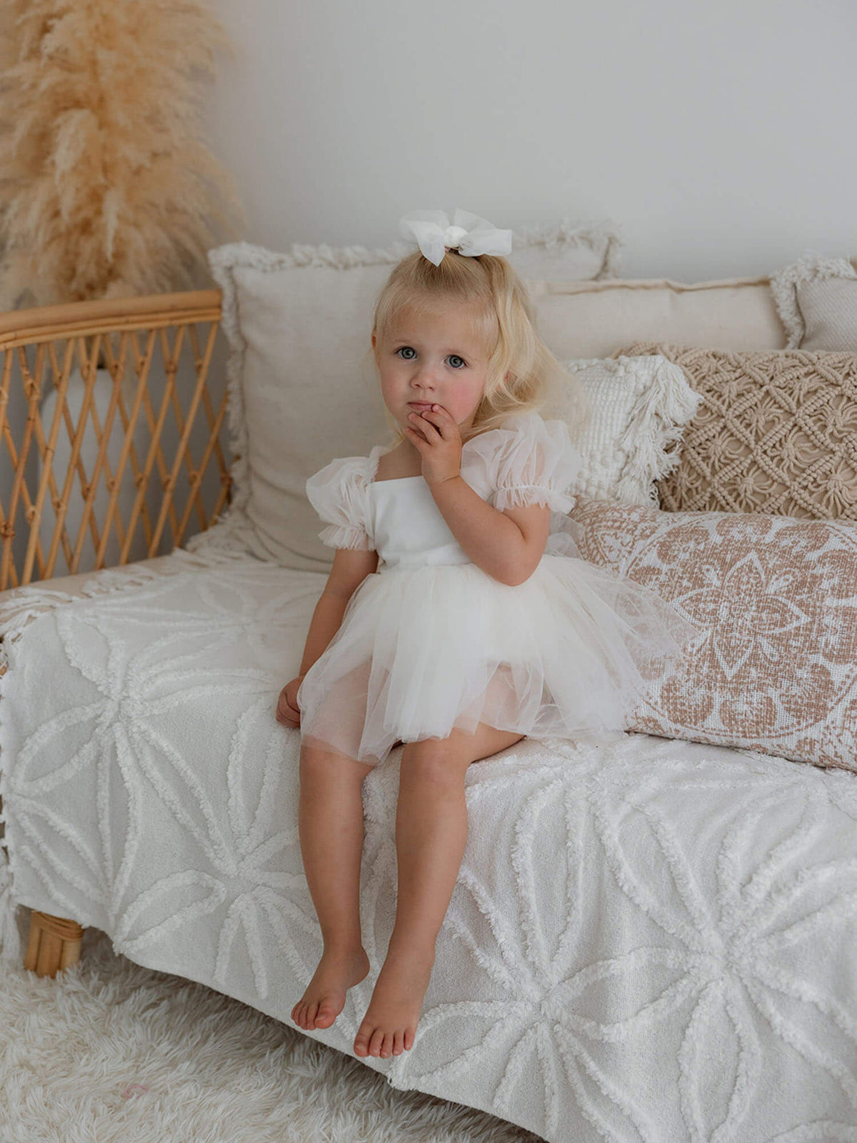 A toddler sits wearing our cream Gabrielle flower girl romper, she also wears a cream tulle bow in her hair.