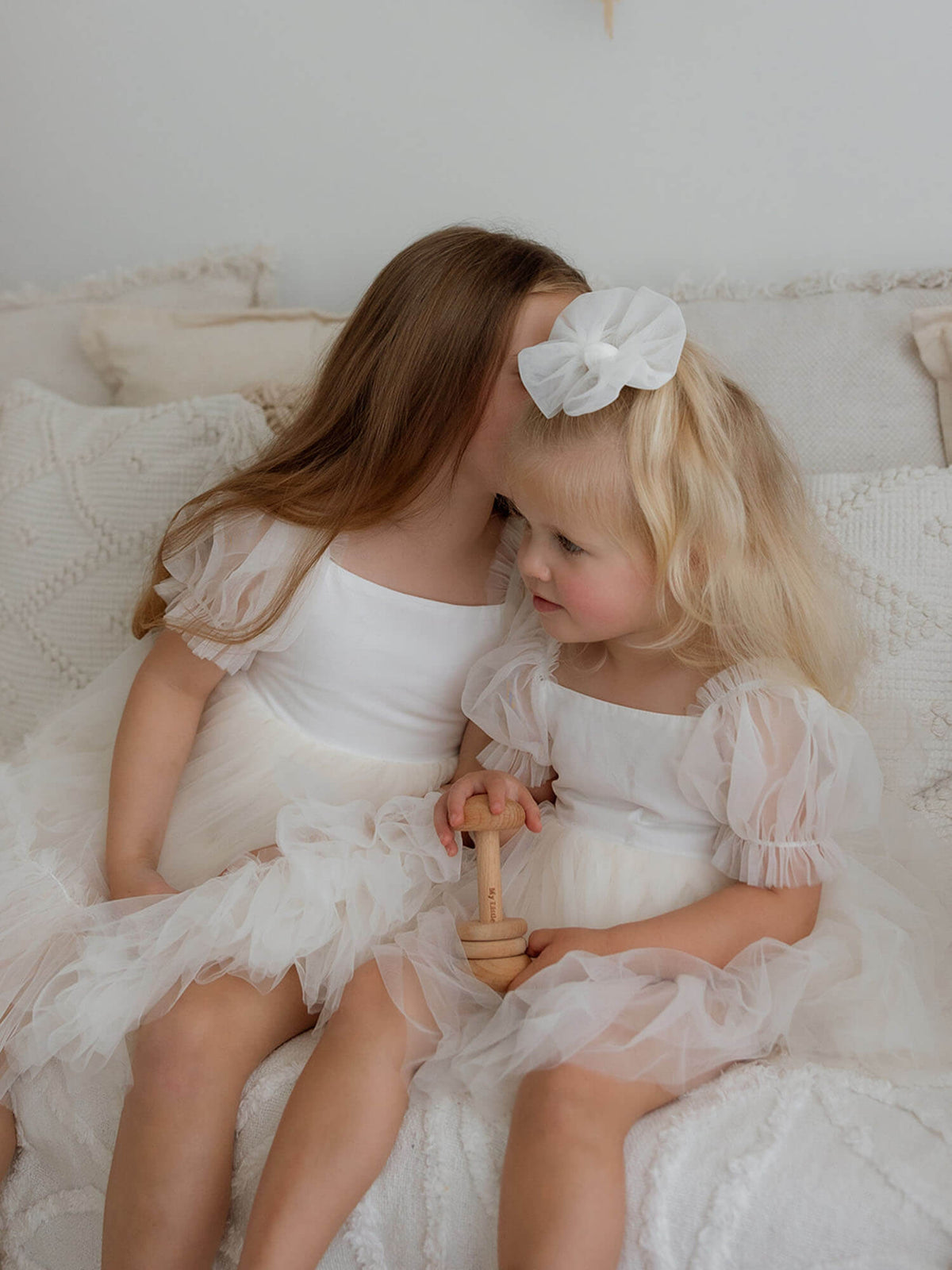 Two young girls sitting together, one wearing our Gabrielle cream flower girl dress and the other in our Gabrielle cream baby flower girl romper with a matching tulle hair bow.