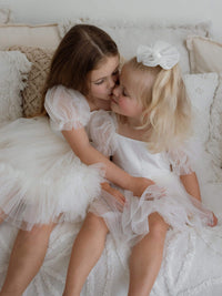 A young girl kisses her sister on the cheek, dressed in matching Gabrielle cream flower girl dress and Gabrielle cream baby flower girl romper.