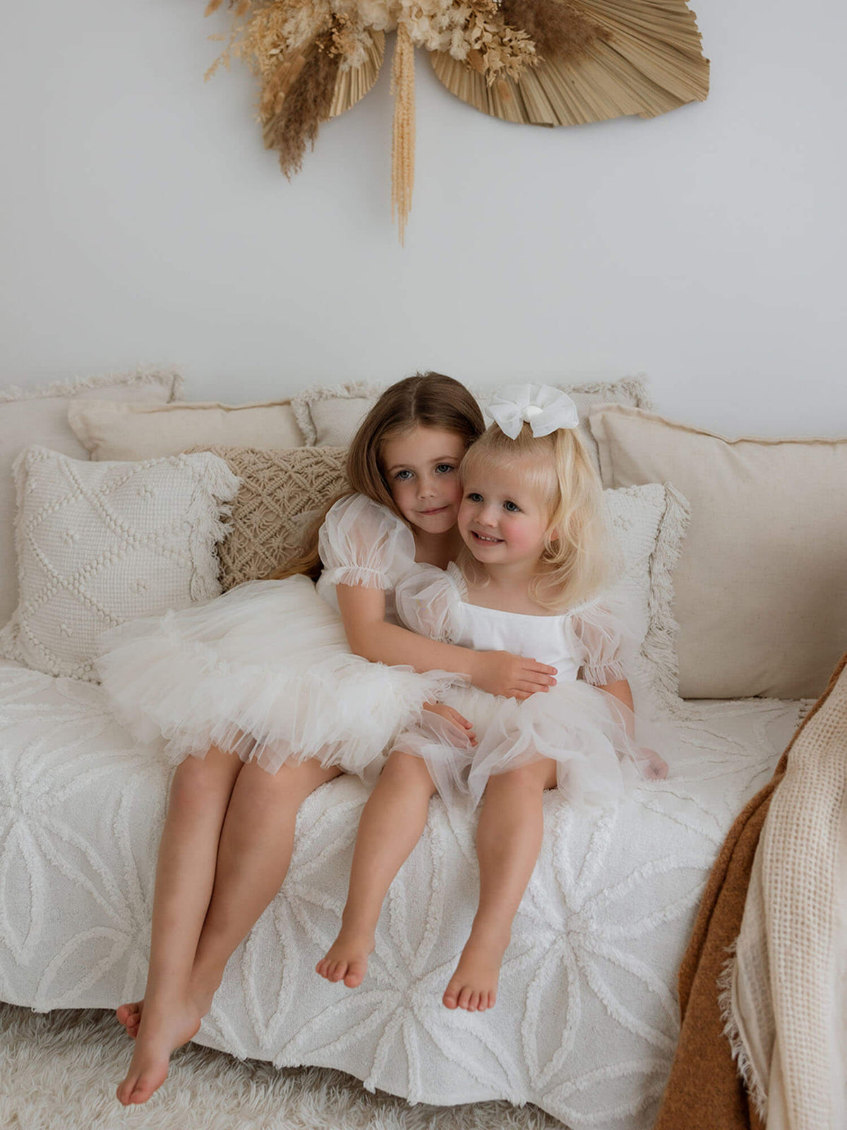 Sisters sit wearing matching flower girl outfits. One sister wears our cream Gabrielle flower girl dress and her younger sister wears our cream Gabrielle baby flower girl romper.