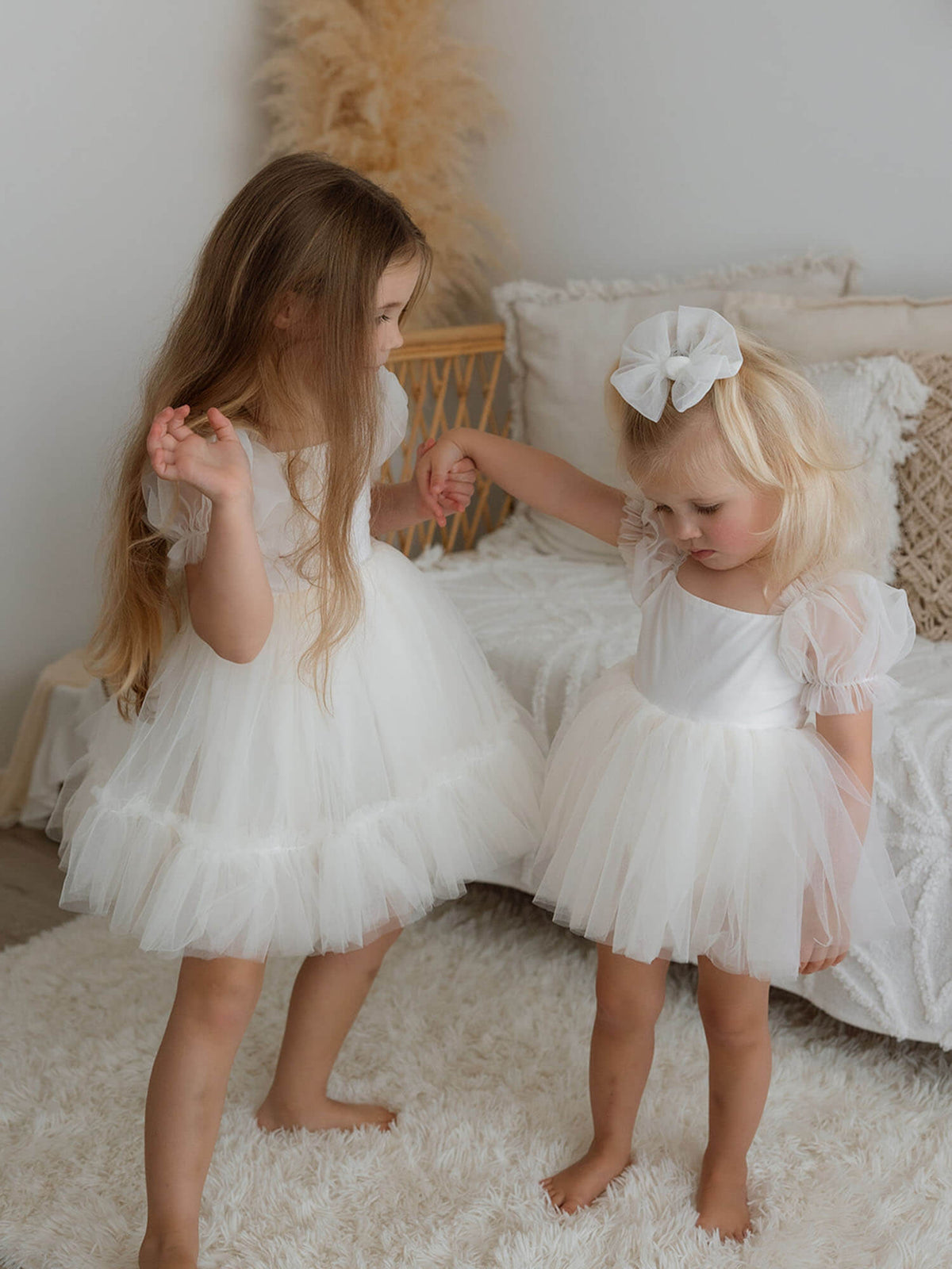 Two girls holding hands, one in our Gabrielle cream flower girl dress and the other in a cream Gabrielle baby flower girl romper with a matching tulle hair bow.